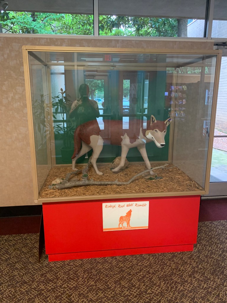 Ceramic wolf sculpture in a glass case indoors. The plaque below reads "Raleigh Red Wolf Ramble"