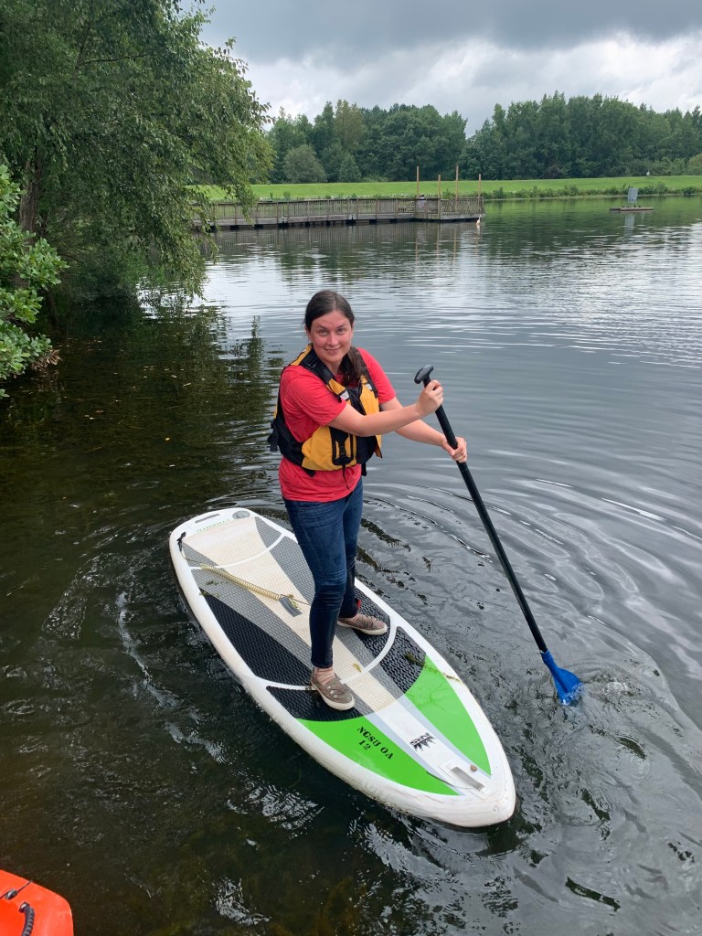 The author standing on a paddleboard on Lake Raleigh, dipping an oar into the water. Trees and grass in the distance, with a cloudy sky overhead