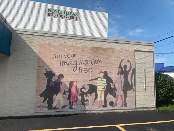 On a building with a sign that reads "Novel Ideas: Used Books - Gifts", a mural is painted that says, "Set your imagination free". The mural includes mundane modern figures with interesting shadows: a schoolboy whose shadow looks like a medieval prince with a feather in his cap, a schoolgirl whose shadow holds a bubbling beaker, a dog whose shadow is shaped like a dragon's, a man in a striped shirt whose shadow is shaped like a soccer player kicking a ball, and a woman in a business outfit whose shadow looks like a ballerina. There is also a cut-off shadow, of which you can only see an arm holding what appears to be a ladel, and part of the torso and one leg. The credits in the corner read, "Lindsay Quick, Milliken University, 2015."